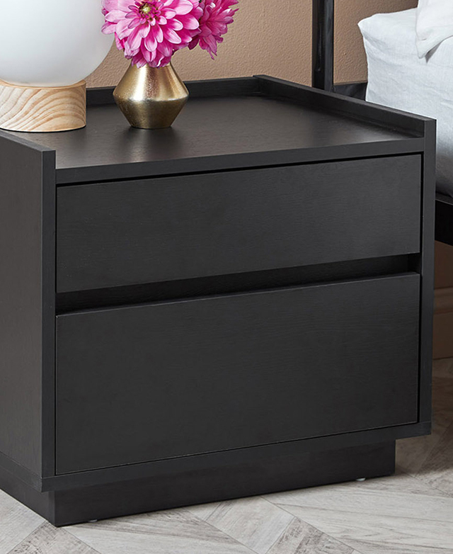 The wide black bedside table is positioned next to a bed, and features pink flowers and a simple spherical lamp on top. The wide black bedside table is positioned next to a bed, and features pink flowers and a simple spherical lamp on top.
