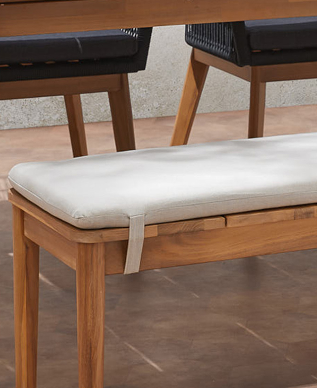 Dappled light plays over a grey bench cushion, which is strapped to the acacia wood base. Dappled light plays over a grey bench cushion, which is strapped to the acacia wood base.