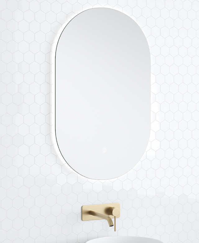 The frameless pill-shaped bathroom mirror is pictured against a white tiled wall above a brass mixer.