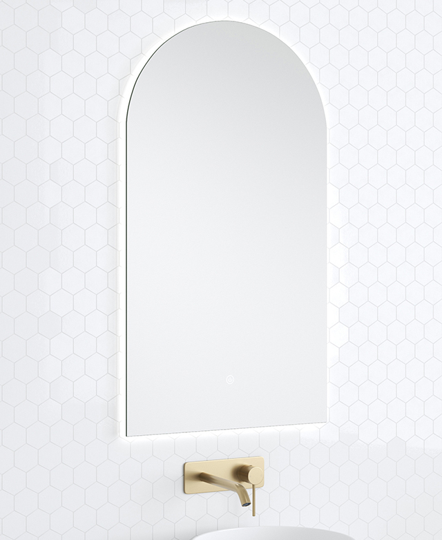The frameless arched bathroom mirror is pictured against a white tiled wall above a brass mixer.