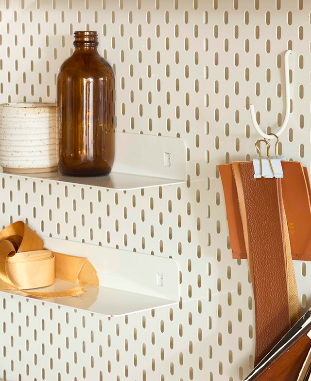 The shelves and hook on the metal pegboard are filled with swatches, a bottle, and a ceramic cup. The shelves and hook on the metal pegboard are filled with swatches, a bottle, and a ceramic cup.