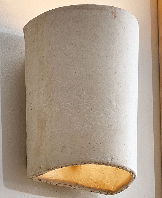 Soft light is seen at the bottom and the top of the cast stone wall sconce, which is placed on a white wall.
