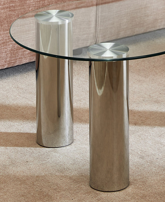 On top of a clay flat weave rug, two of the polished chrome pillar legs support the clear glass coffee table top.
