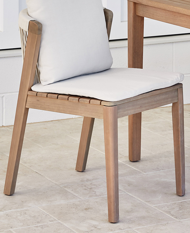 A dining chair topped with white cushions is positioned next to a table on a cool, tiled outdoor floor.