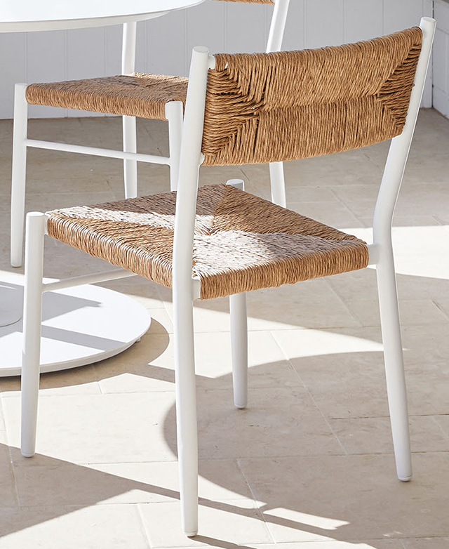 A back view of the wicker and metal outdoor dining chair is positioned next to a white table on a terrace.