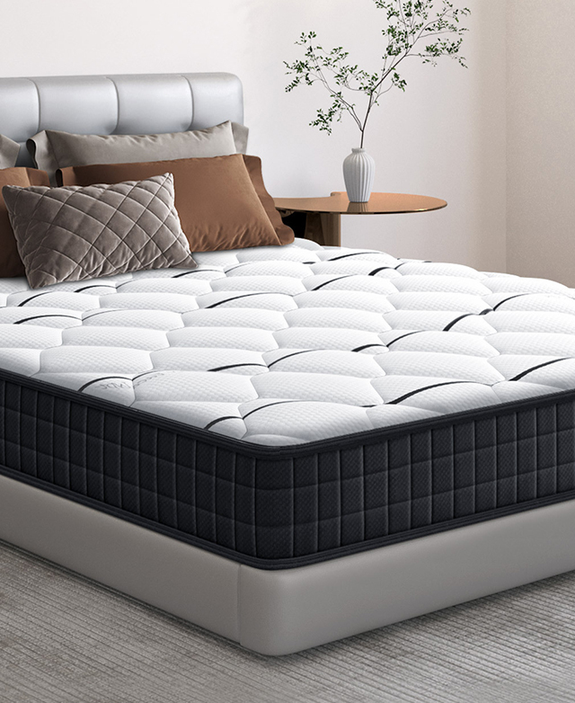 The mattress is on a grey bed, which has a foliage-topped bedside table next to it; several brown-toned pillows are on top. The mattress is on a grey bed, which has a foliage-topped bedside table next to it; several brown-toned pillows are on top.