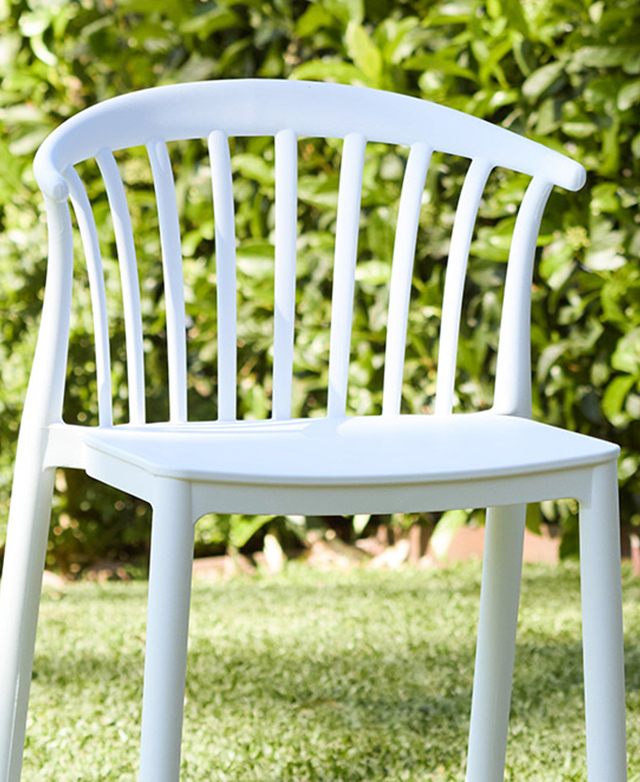 The white bar stool is shown in an outdoor setting on grass with a verdant hedge in the background.
