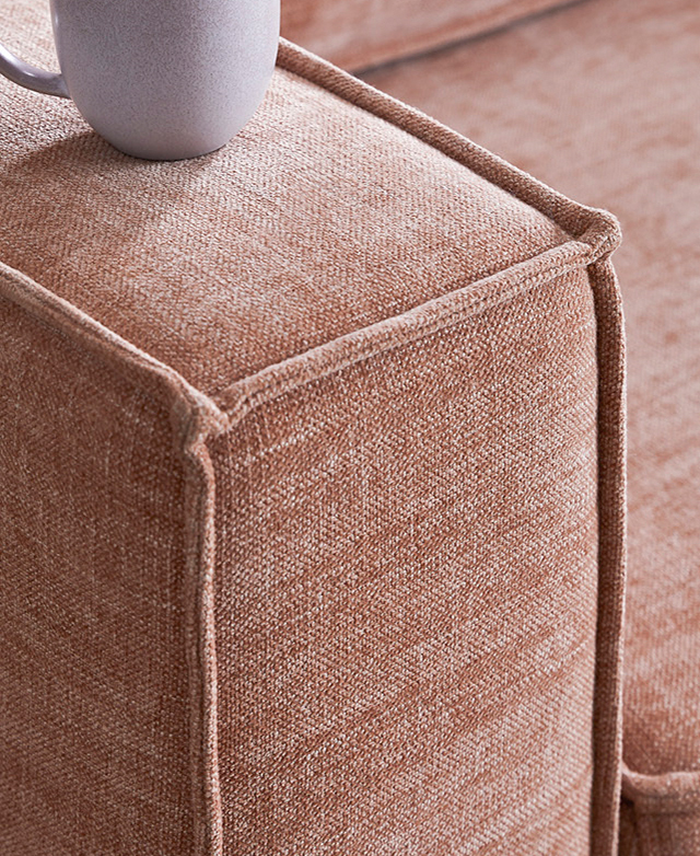 The blush sofa is zoomed in on to show a cream ceramic mug on the thick armrest, and the neat piping trims.