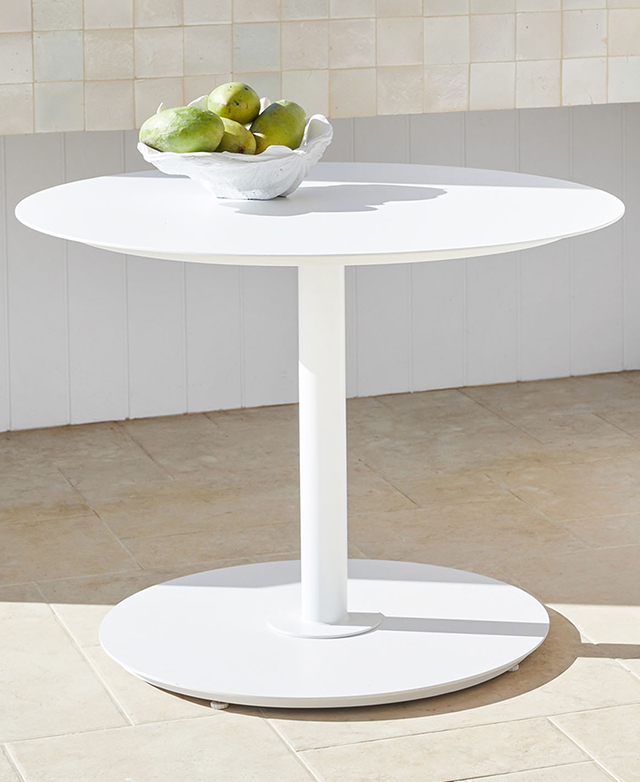 The round white metal outdoor table is depicted in sunlight on a stone tiled floor, with a bowl of fruit on top. The round white metal outdoor table is depicted in sunlight on a stone tiled floor, with a bowl of fruit on top.