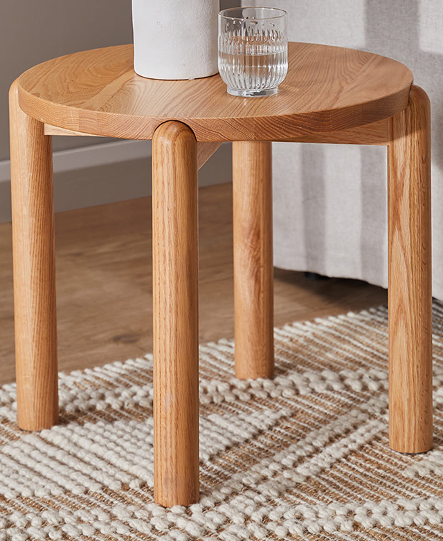 A natural oak wood round side table is on a jute and wool rug next to a chair, with a glass of water and ceramic on top. A natural oak wood round side table is on a jute and wool rug next to a chair, with a glass of water and ceramic on top.