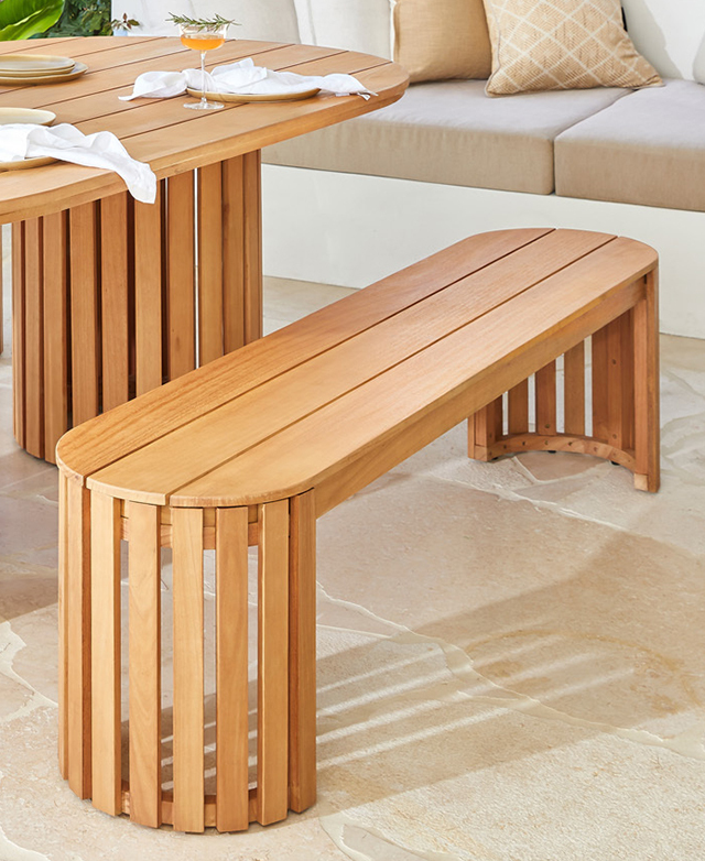 A long, oval-shaped eucalyptus wood outdoor bench is positioned next to a dining table on a stone floor.