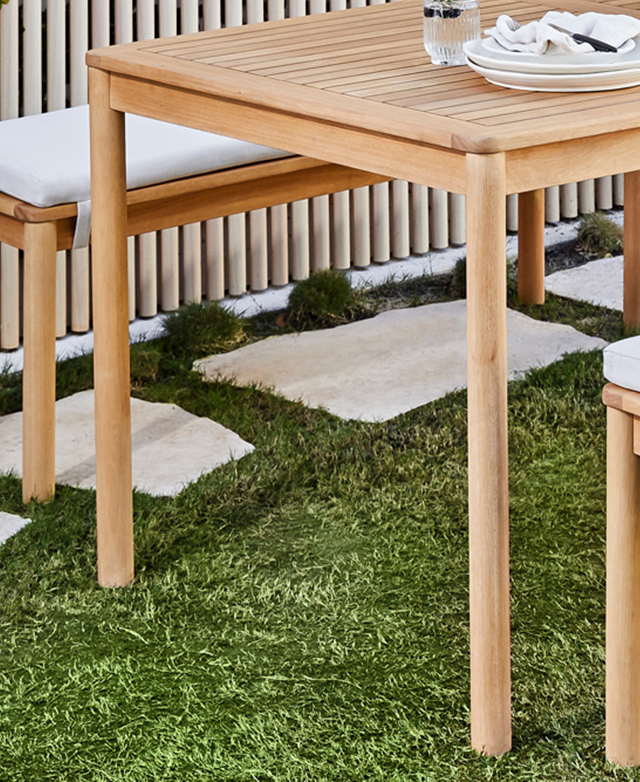 A dining table and cushioned bench set are on grass with stepping stones, with crockery and napery on the table top. A dining table and cushioned bench set are on grass with stepping stones, with crockery and napery on the table top.