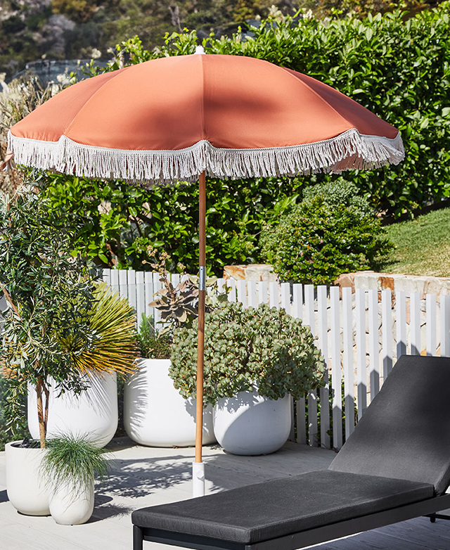 The orange umbrella with white fringing is erected outside next to a white picket fence and a black cushioned sun lounger. The orange umbrella with white fringing is erected outside next to a white picket fence and a black cushioned sun lounger.