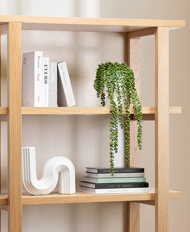The top tiers of the bookshelf are adorned with stacks of books, a white abstract ceramic, and a draping plant.