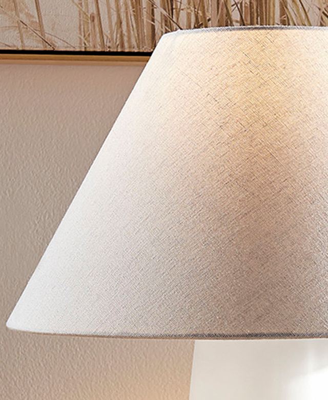 The off-white empire shade is illuminated by the hidden bulb; in the background is a pampas grass print on the wall.