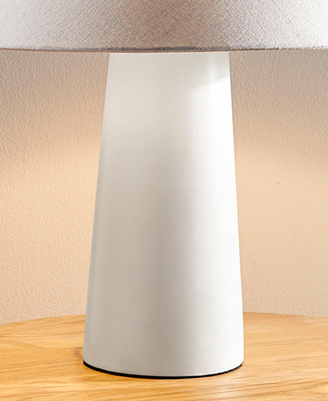 The simple, white, pillar ceramic lamp base is illuminated and on a timber surface.