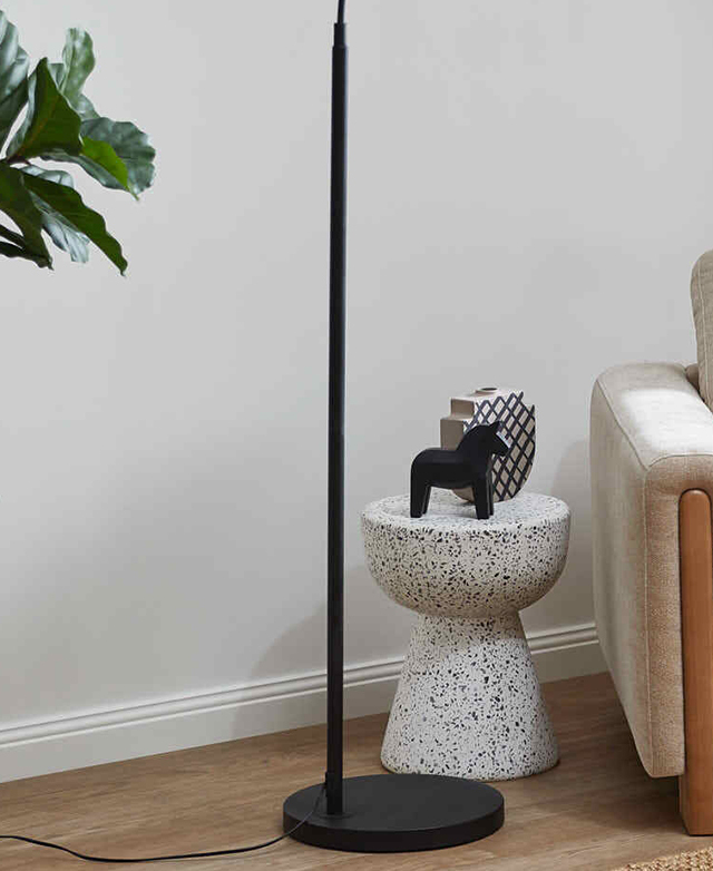 The black steel, slender lamp base is on a timber floor next to a terrazzo side table and a neutral armchair. The black steel, slender lamp base is on a timber floor next to a terrazzo side table and a neutral armchair.