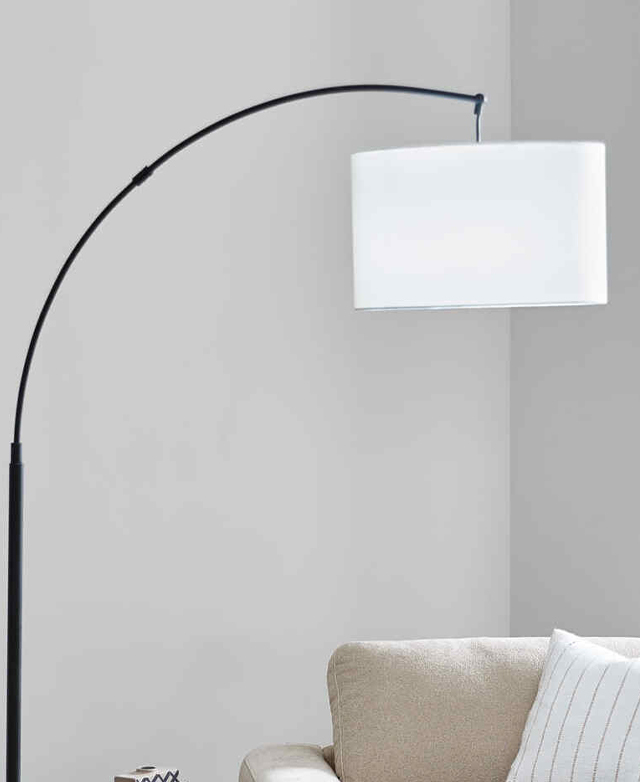 The white fabric drum shade hangs as if on the end of a fish hook in front of a white wall, over a neutral armchair. The white fabric drum shade hangs as if on the end of a fish hook in front of a white wall, over a neutral armchair.