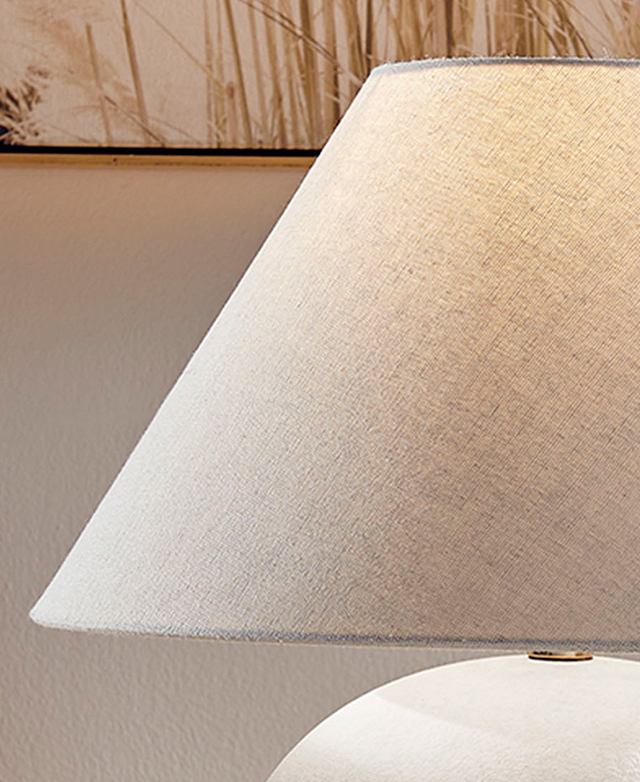The white empire shade is illuminated by the hidden bulb; in the background is a pampas grass print on the wall.