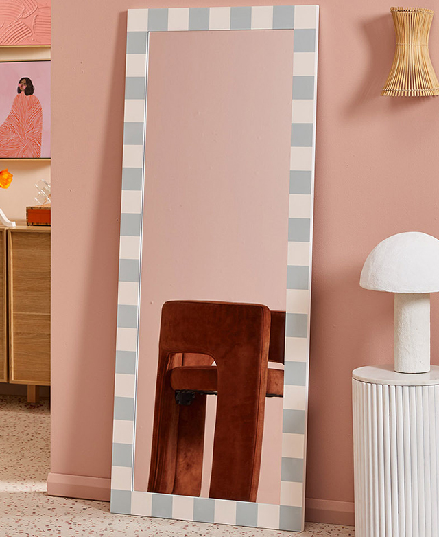 The checkered full length mirror leans against a soft pink wall and reflects a rust, velvet armchair.