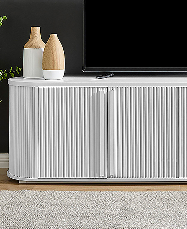 The white entertainment unit is in front of a dark grey wall with a TV on top. The fluted cupboards are on full display.