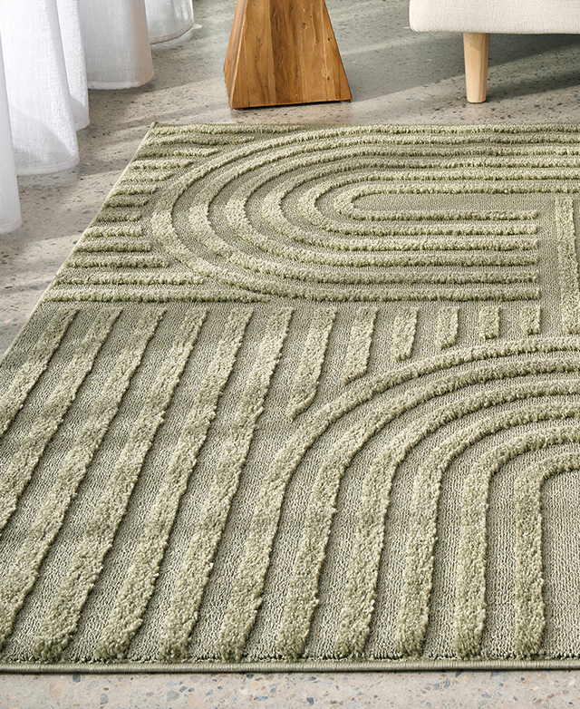 The pistachio rug is laid out on a speckled concrete floor. The high-low cutwork texture is tonal greens.
