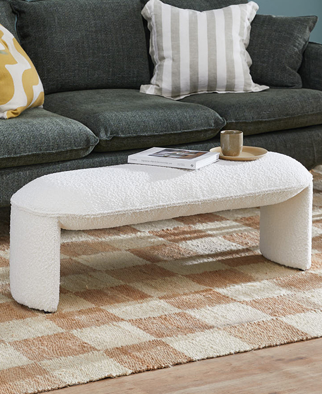 The long creamy ottoman is in front of a green sofa on a taupe & cream checkered rug. A cup, saucer & magazine are on top. The long creamy ottoman is in front of a green sofa on a taupe & cream checkered rug. A cup, saucer & magazine are on top.