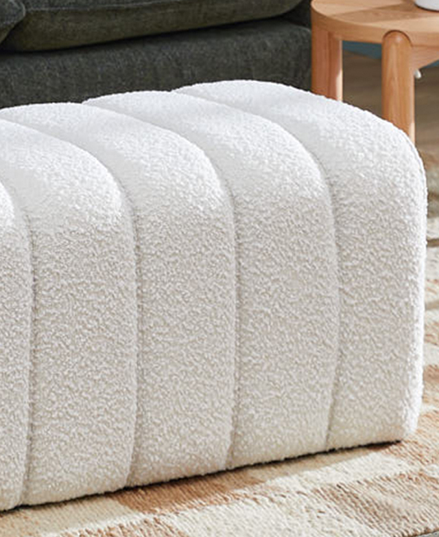 Cropped view of the textured boucle ottoman shows one end in a living room on top of a taupe and cream checkered rug. Cropped view of the textured boucle ottoman shows one end in a living room on top of a taupe and cream checkered rug.