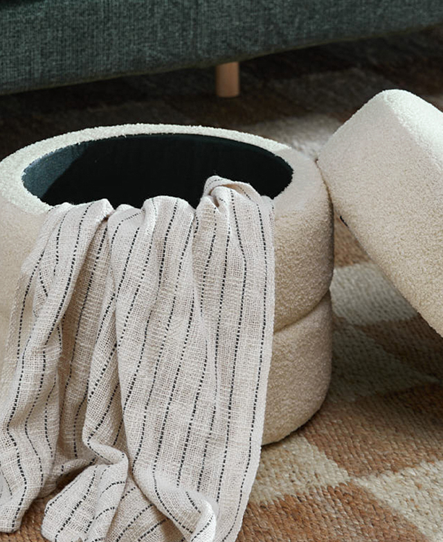 The cream storage ottoman is shown with its lid off, with a monochrome stripey blanket spilling out of it. The cream storage ottoman is shown with its lid off, with a monochrome stripey blanket spilling out of it.
