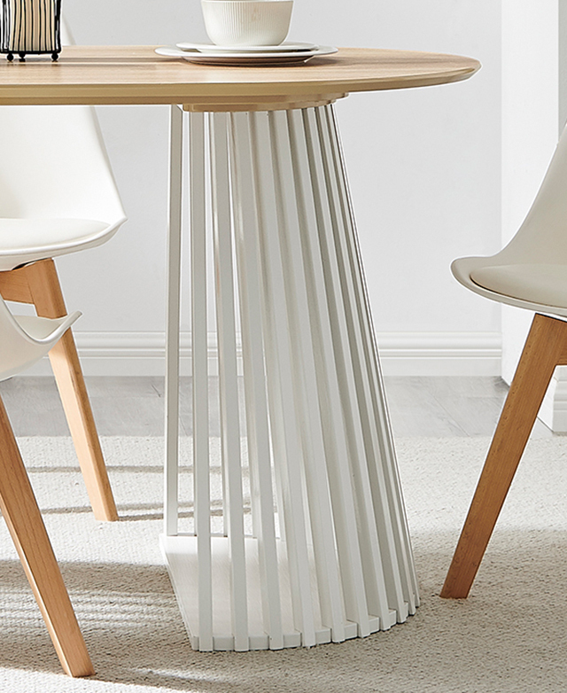 One strutted pillar end of the white and oak table is shown on top of a cream rug. Dining chairs are around it. One strutted pillar end of the white and oak table is shown on top of a cream rug. Dining chairs are around it.