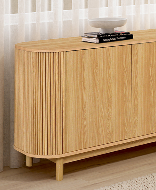 Three-quarter angle highlights the smooth cupboard doors and the fluted, rounded end. It's in front of a cream mesh curtain.