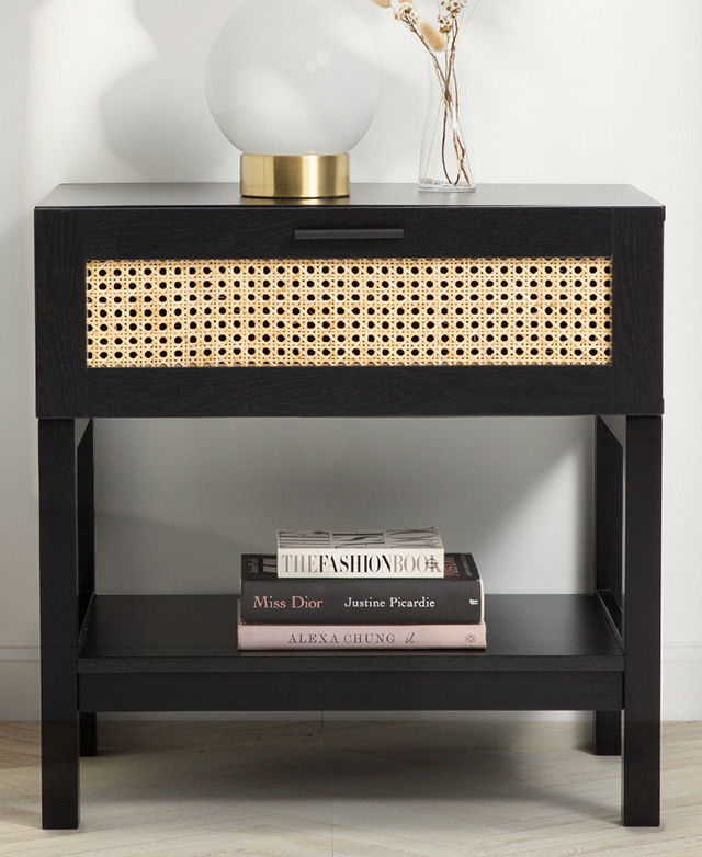 A black table is positioned in front of a white wall, the drawer's rattan panel is the focus. Decor is styled on top.