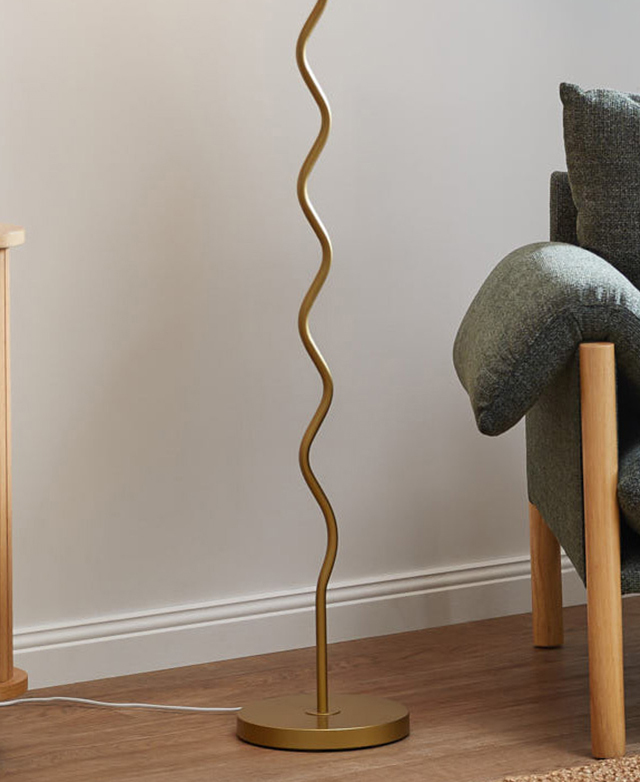 A golden metal lamp base is wavy with a round stand. It's on a timber floor in a white room, next to an armchair. A golden metal lamp base is wavy with a round stand. It's on a timber floor in a white room, next to an armchair.