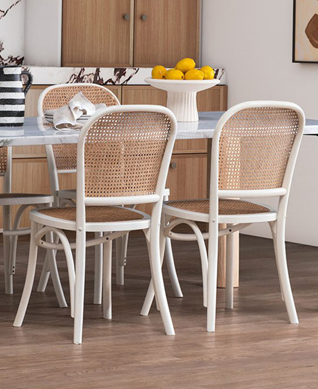 Two white and natural rattan dining chairs are shown from behind, pulled up to a dining table in a bright dining room.