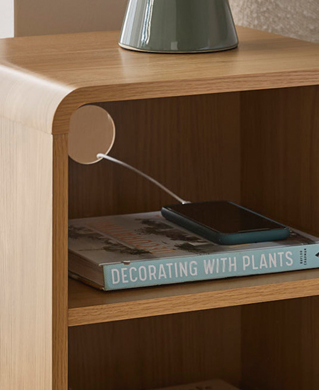The top shelf has a book on it, with a phone on top that's attached to a charging cord, which is fed through the cable hole. The top shelf has a book on it, with a phone on top that's attached to a charging cord, which is fed through the cable hole.