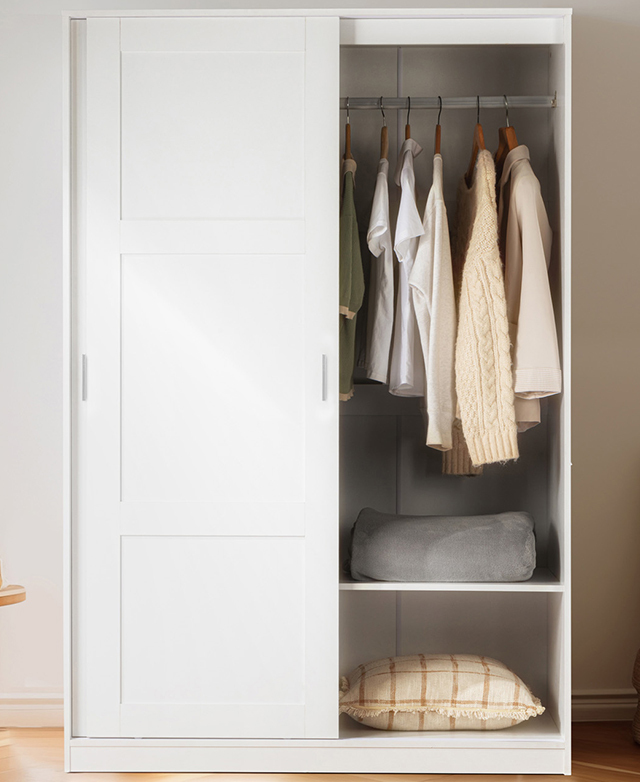 The full white wardrobe's sliding door is open to reveal hung clothing and folded bedding. The full white wardrobe's sliding door is open to reveal hung clothing and folded bedding.