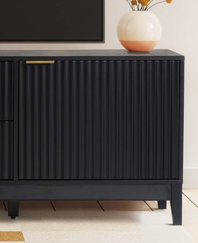 One end of the black TV unit against an off-white wall, highlighting the fluted doors and a brass tab handle. One end of the black TV unit against an off-white wall, highlighting the fluted doors and a brass tab handle.