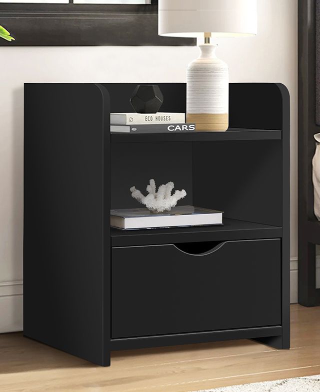 The black bedside table is adorned with a cream lamp, books, and decor; it's positioned against a white wall.