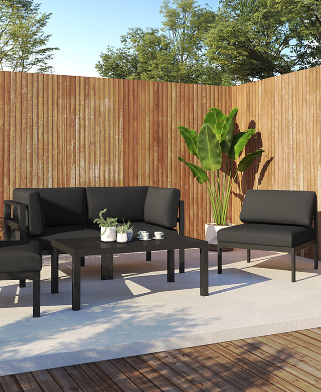 Framed by timber panelled walls and banana leaves, the dark grey outdoor lounge set is arranged around a coffee table.