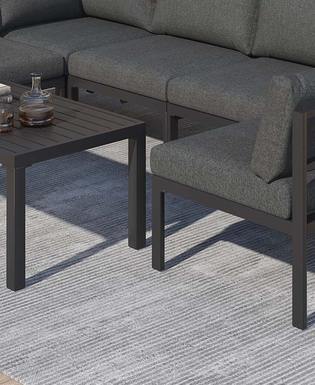 Cropped view shows the charcoal grey outdoor lounge positioned around a metal coffee table on a grey rug. Cropped view shows the charcoal grey outdoor lounge positioned around a metal coffee table on a grey rug.