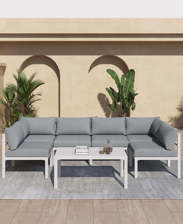A bright, sunlit outdoor area, complete with stone backdrop & leafy plants, holds a grey &white lounge & coffee table set. A bright, sunlit outdoor area, complete with stone backdrop & leafy plants, holds a grey &white lounge & coffee table set.