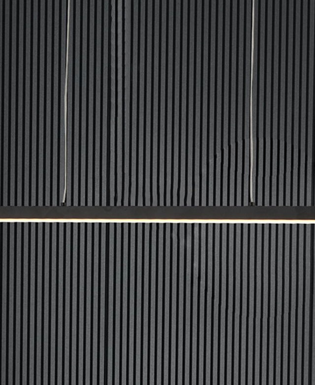 The black bar pendant light with the light on is in front of a charcoal panelled wall.