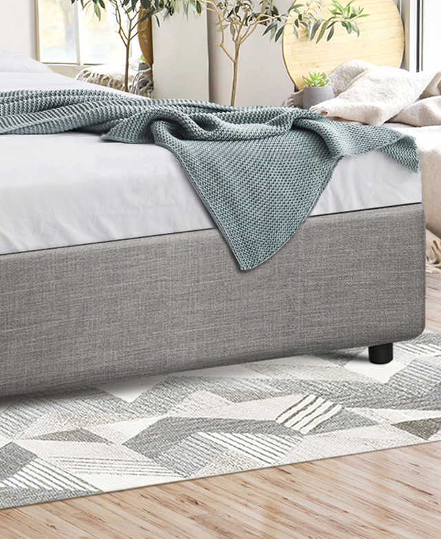 The end of the grey upholstered bed with mattress is atop a geomteric monochrome rug, & a blue throw is draped over the top. The end of the grey upholstered bed with mattress is atop a geomteric monochrome rug, & a blue throw is draped over the top.