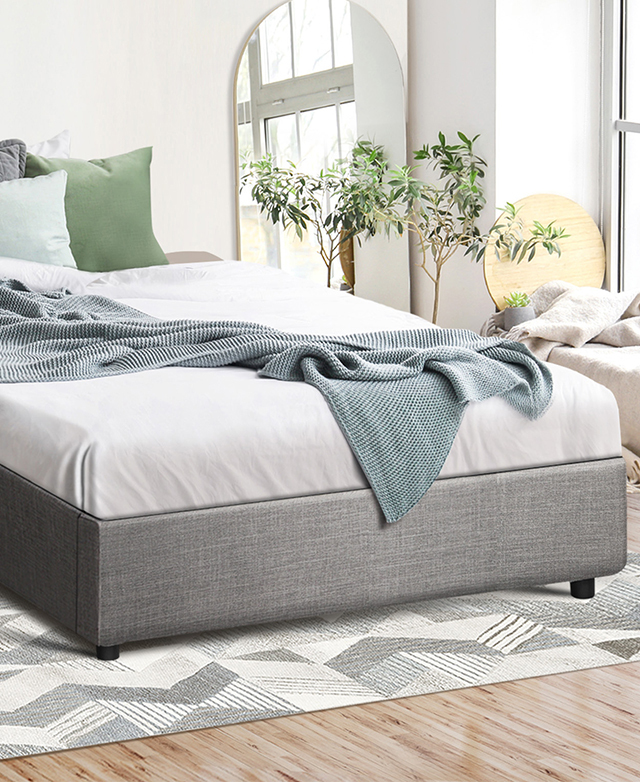 The grey upholstered bed is dressed with white sheets and a blue blanket in a bright room. The grey upholstered bed is dressed with white sheets and a blue blanket in a bright room.