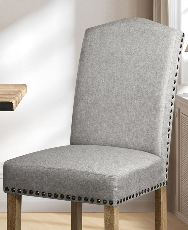 A dining chair is in a bright room; the grey upholstery of the seat and back is trimmed with metal studwork. A dining chair is in a bright room; the grey upholstery of the seat and back is trimmed with metal studwork.