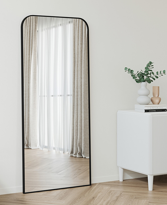 Oversized black-framed mirror leans against a white wall, on parquet timber flooring next to a white console. Oversized black-framed mirror leans against a white wall, on parquet timber flooring next to a white console.