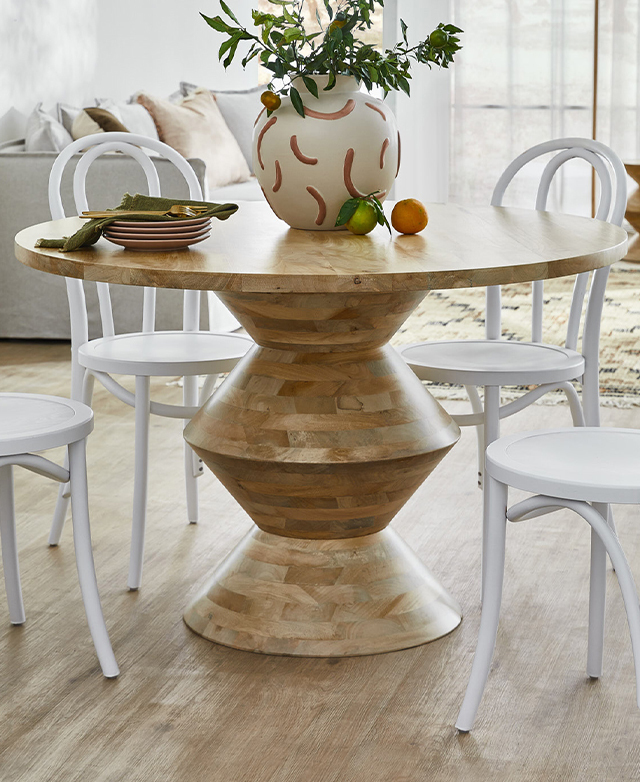 The round pedestal dining table is styled with white Thonet chairs, with crockery and a large vase filled with leaves on top. The round pedestal dining table is styled with white Thonet chairs, with crockery and a large vase filled with leaves on top.