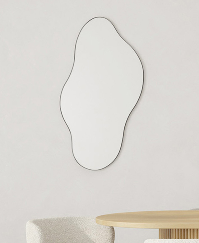 Minimalistic styling places the narrow-framed wavy mirror on a white wall above a table and chair. Minimalistic styling places the narrow-framed wavy mirror on a white wall above a table and chair.