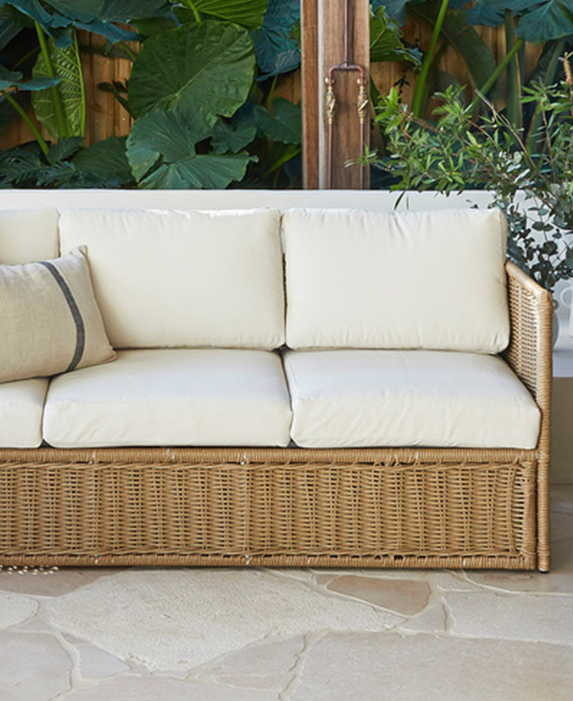 An outdoor sofa features cream seat and back cushions and a polyurethane rattan frame. It has a backdrop of verdant leaves.