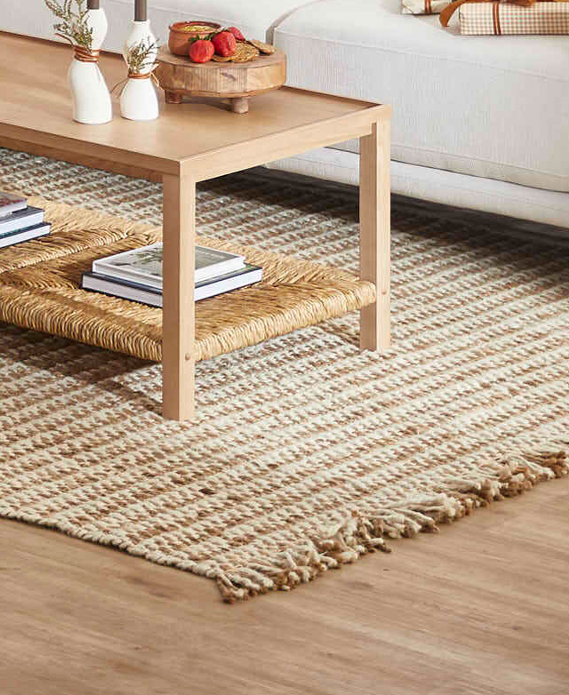 A fringed, natural jute rug is positioned in a living room next to a cream sofa, under a timber and rush coffee table. A fringed, natural jute rug is positioned in a living room next to a cream sofa, under a timber and rush coffee table.
